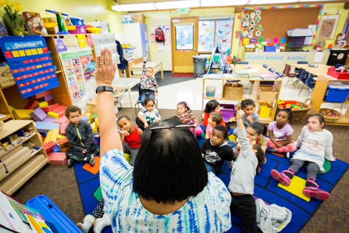 Pre-school teacher in a colorful classroom raises her hand, young children look up to her